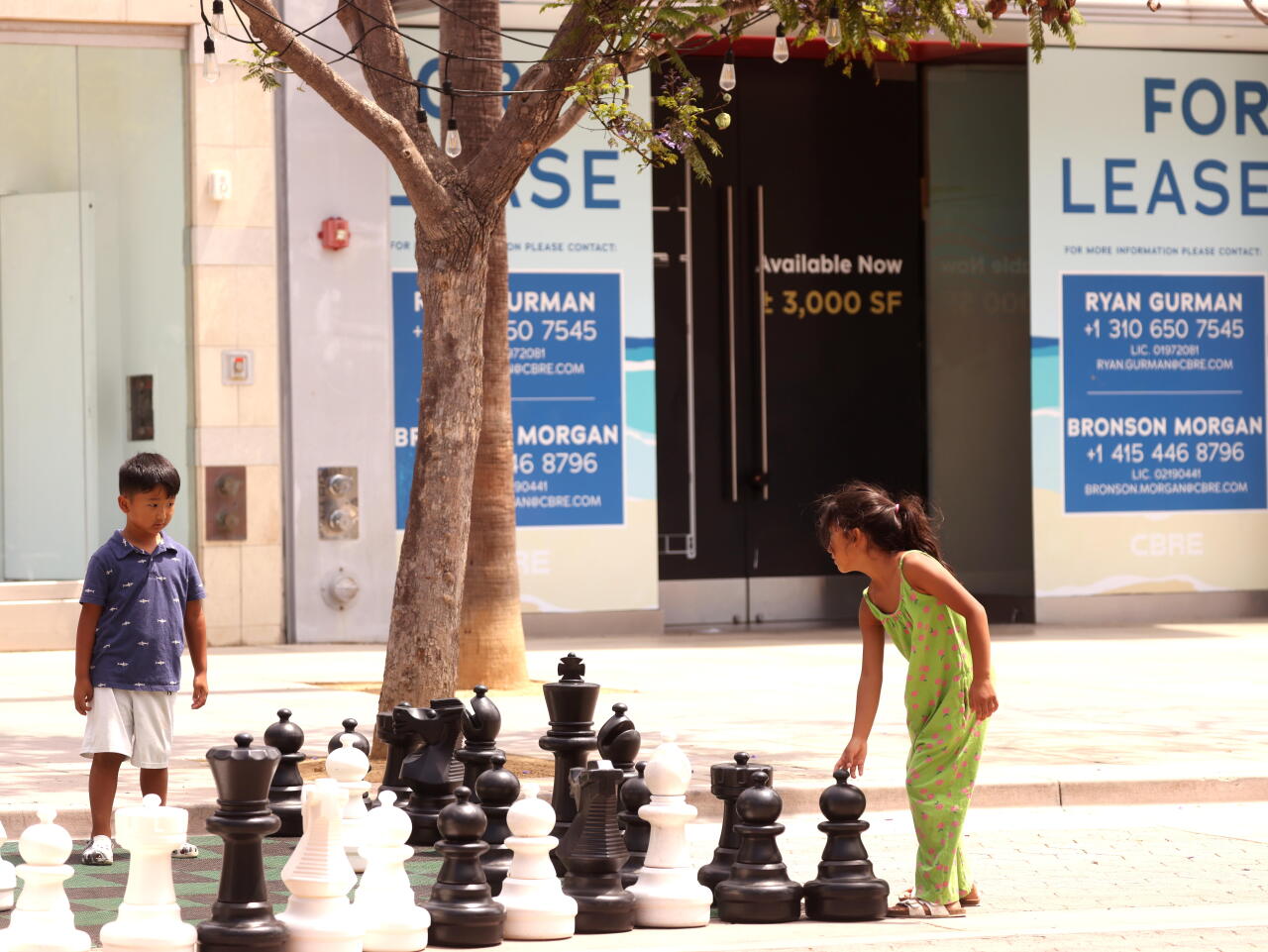 Two children stand next to giant chess pieces on the ground.