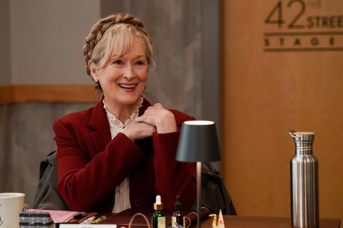 Loretta (Meryl Streep) in a maroon blazer and white shirt sits at a table with a small black lamp.