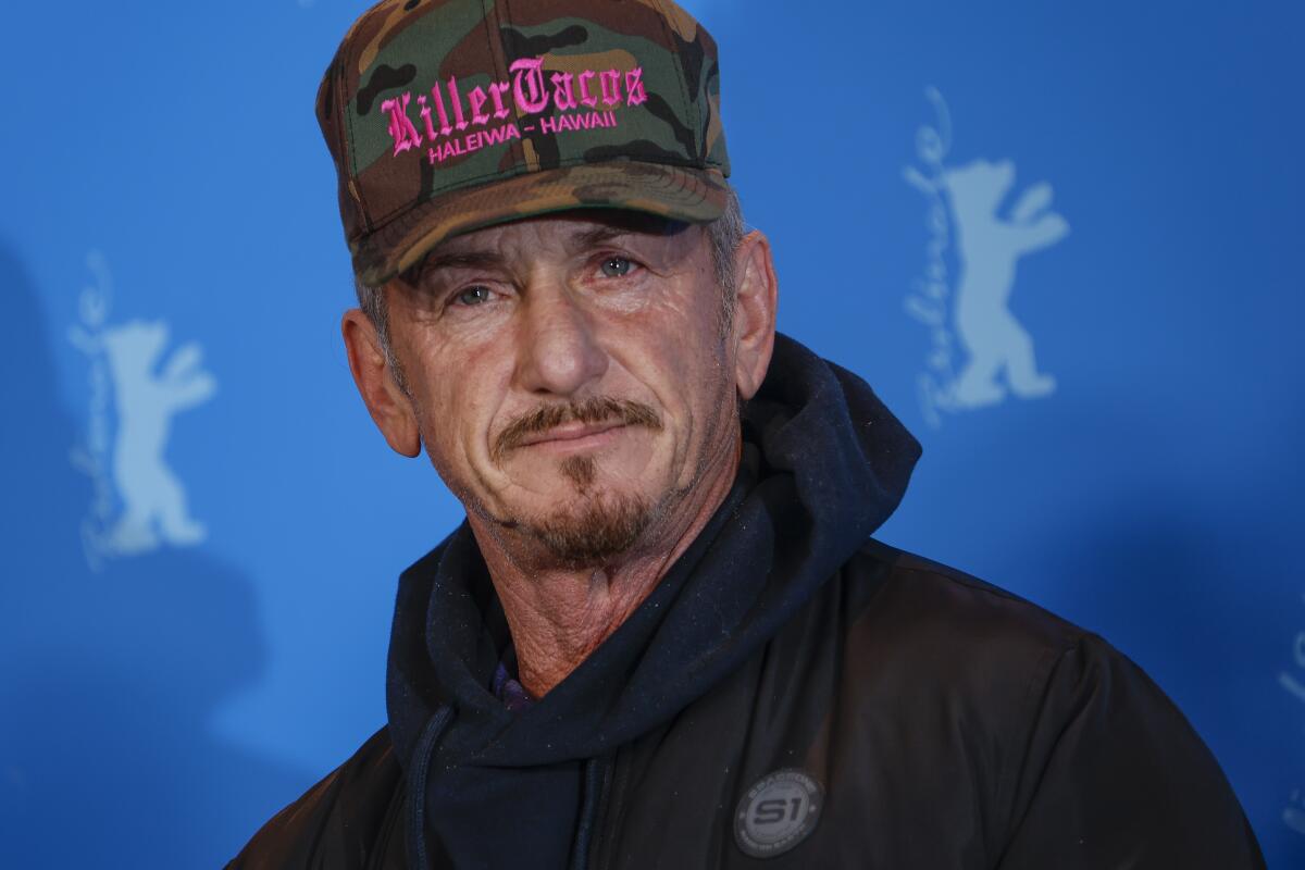Sean Penn wears a camouflage baseball cap and hooded sweatshirt against a blue backdrop.  