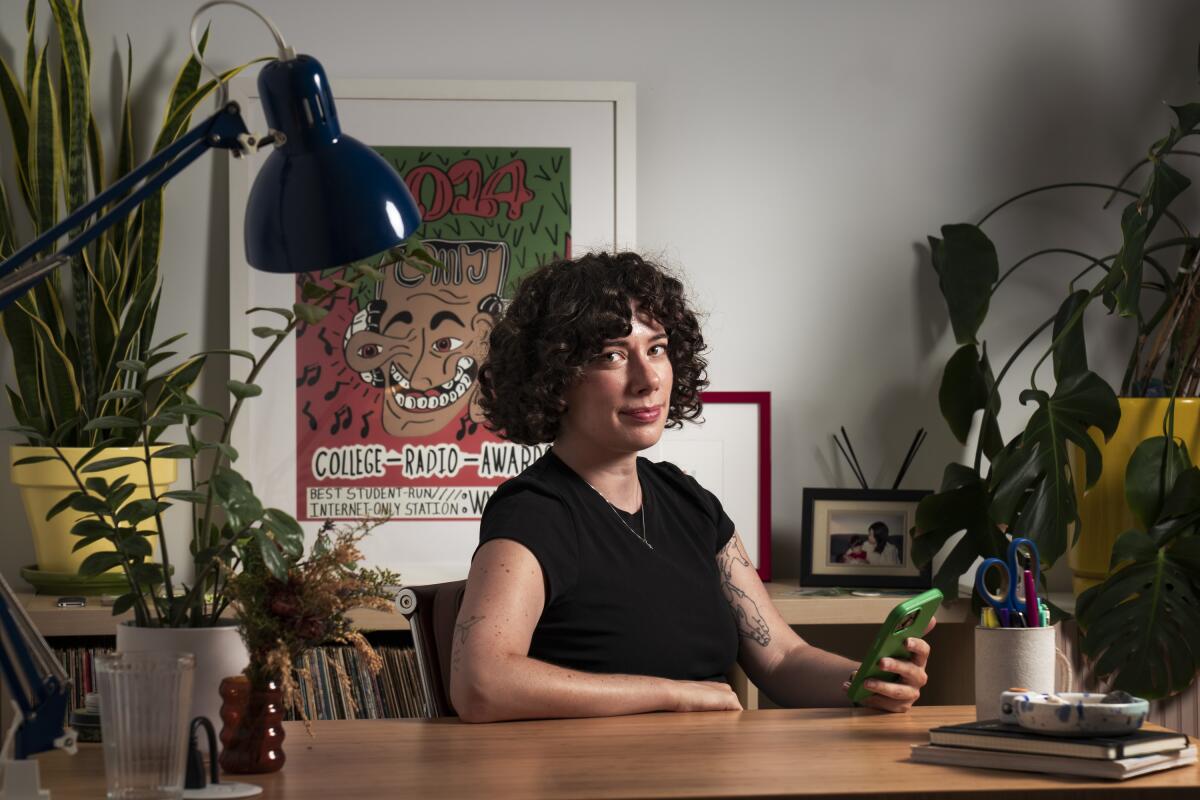 A woman sits at a desk, holding a cellphone, with potted plants around her and a colorful poster on the wall behind her