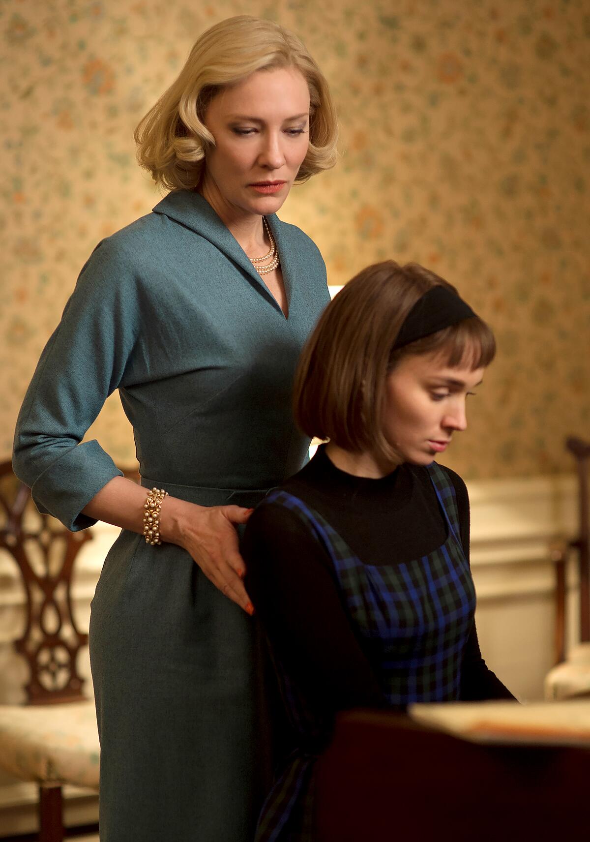 An older woman stands behind a seated younger woman, hands on her shoulders in "Carol."