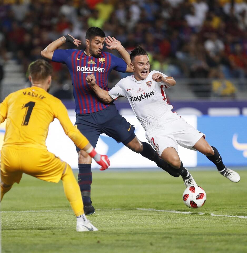 El centrocampista del Sevilla Roque Mesa (d) cae ante el barcelonista Luis Suárez (c), en presencia del portero Vaclik, durante el partido de la Supercopa de España que disputan esta noche en el estadio Ibn Battuta de Tánger, en Marruecos.