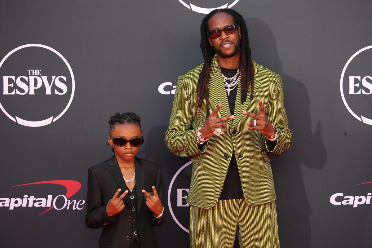 2 Chainz gives the peace sign with both hands while standing alongside a child doing the same.