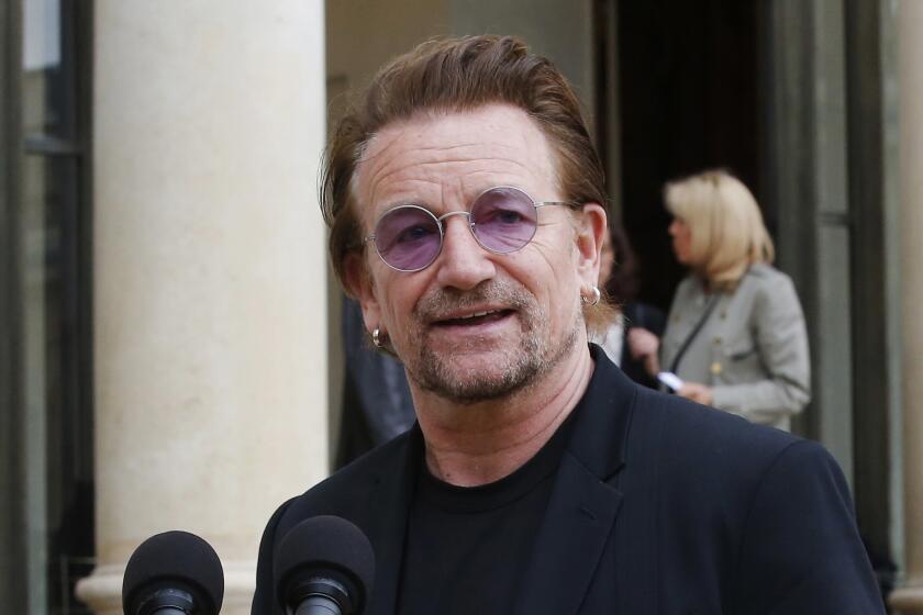 A man wearing purple-hued sunglasses and dark shirt and blazer speaks behind a dual microphone