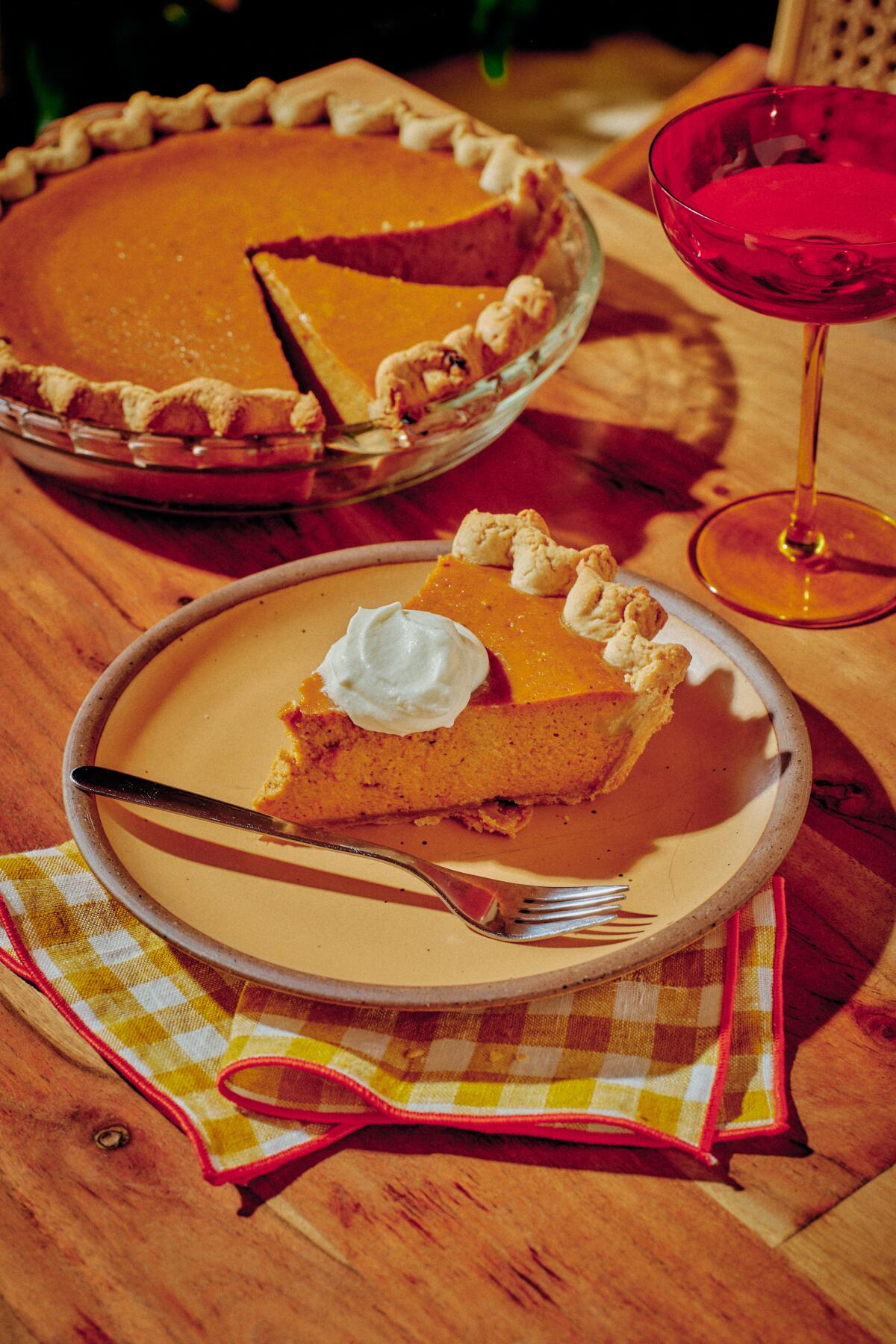 A slice of pumpkin pie with a dollop of whipped cream sits on a plate next to the rest of the whole pie and a glass of wine