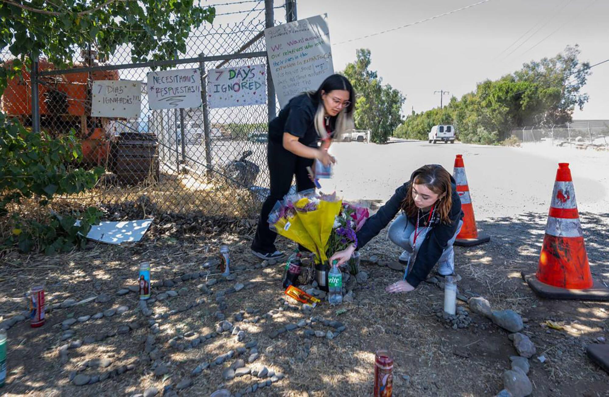 Two young women place items at a memorial site outdoors.