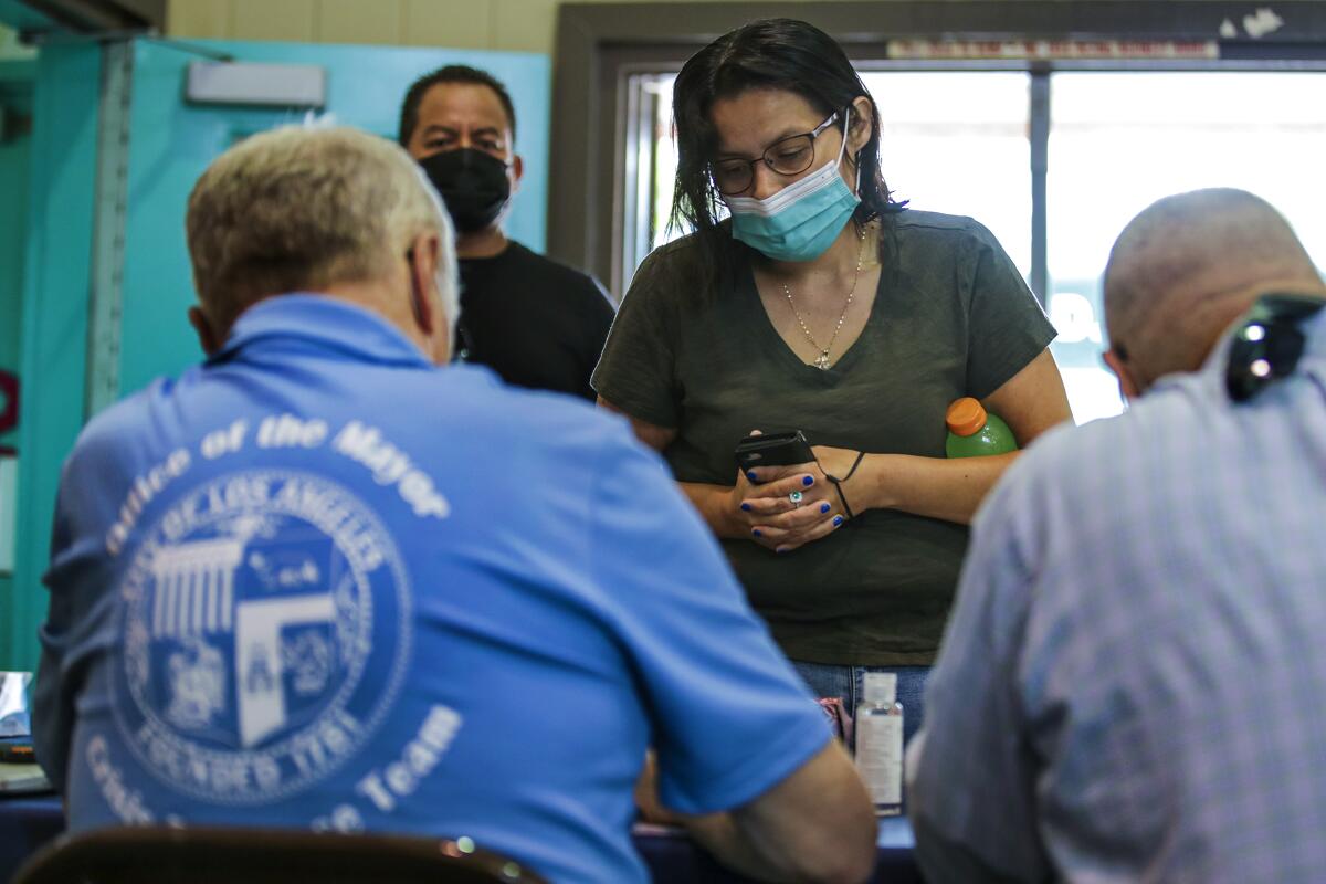 A woman in a mask, with a Gatorade under one arm, clasps her phone as she speaks to two men seated at a table. 