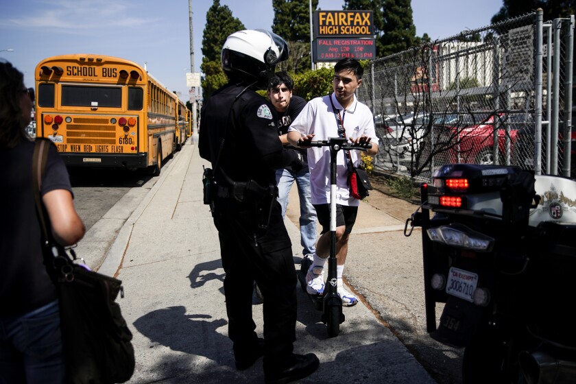 An LAPD bike officer warns two young men about riding their e-scooters on the sidewalk, in front of Fairfax High School near the intersection of Fairfax and Melrose avenues on Thursday.