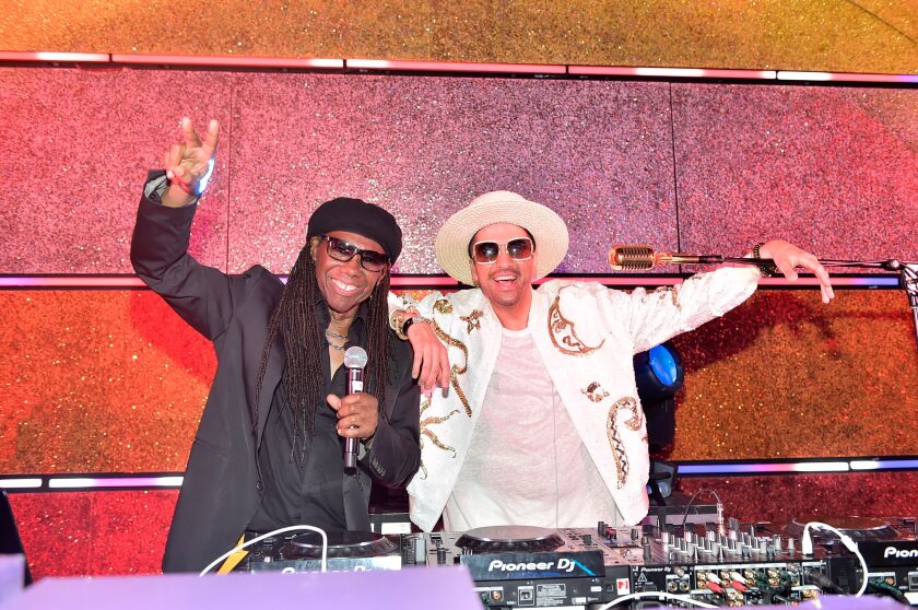 Two people, one holding a microphone and flashing a peace signal, are standing in front of the DJ equipment.
