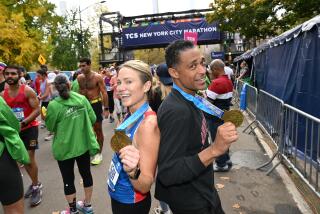 A blond woman with a ponytail in running gear and a Black man in dark running gear posing back to back holding medals