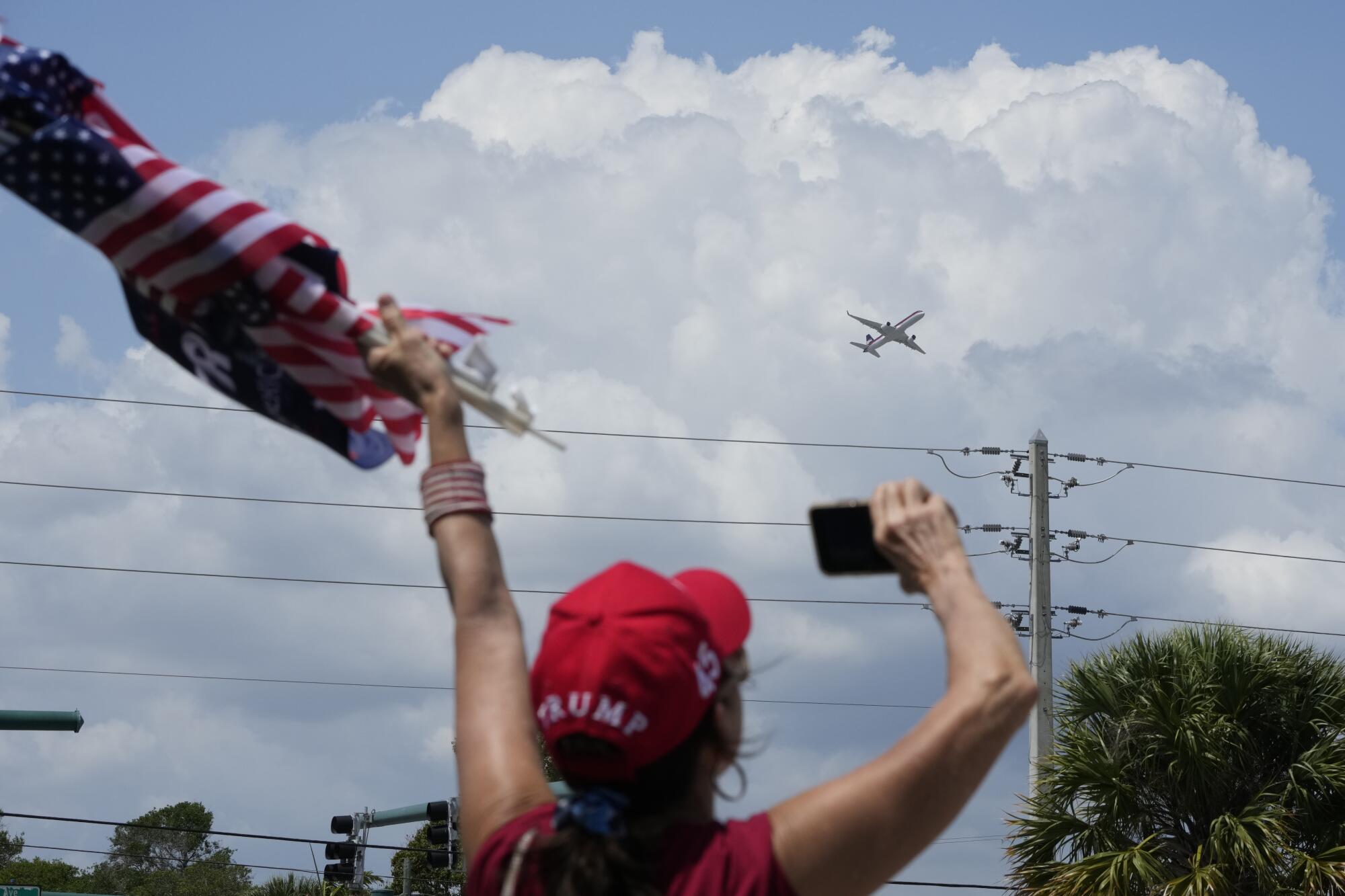 A woman, holding small American flags in her left had and a phone in her right hand, waves toward a plane in sky