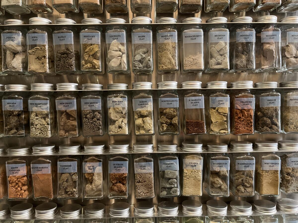 A spice rack is filled with bottles each bearing different colors of earth and geographic labels like "New Mexico Clay."