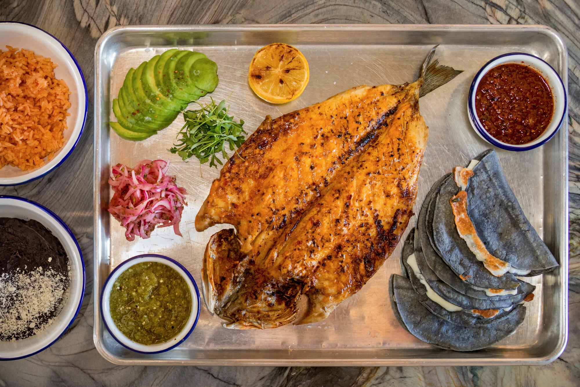 A butterflied cooked fish on a metal tray with tacos and sauces and other toppings.