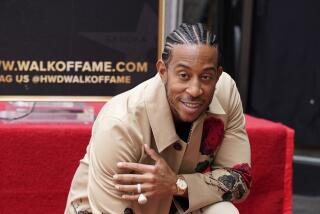 Ludacris, wearing a tan coat and pants, poses with hand resting on his star on the Hollywood Walk of Fame. 