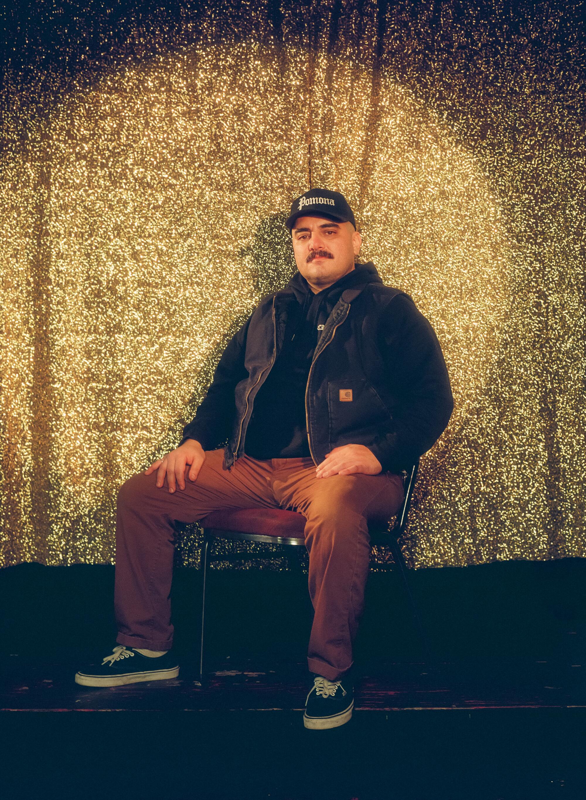 A man in brown pants and a black cap sits in front of a golden curtain.