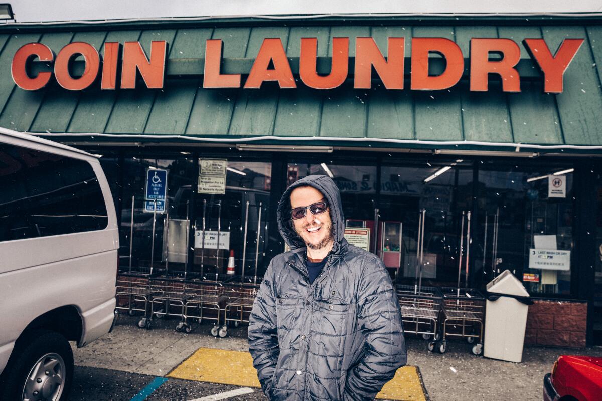 A man wearing a hooded jacket and sunglasses stands in front of a laundromat.