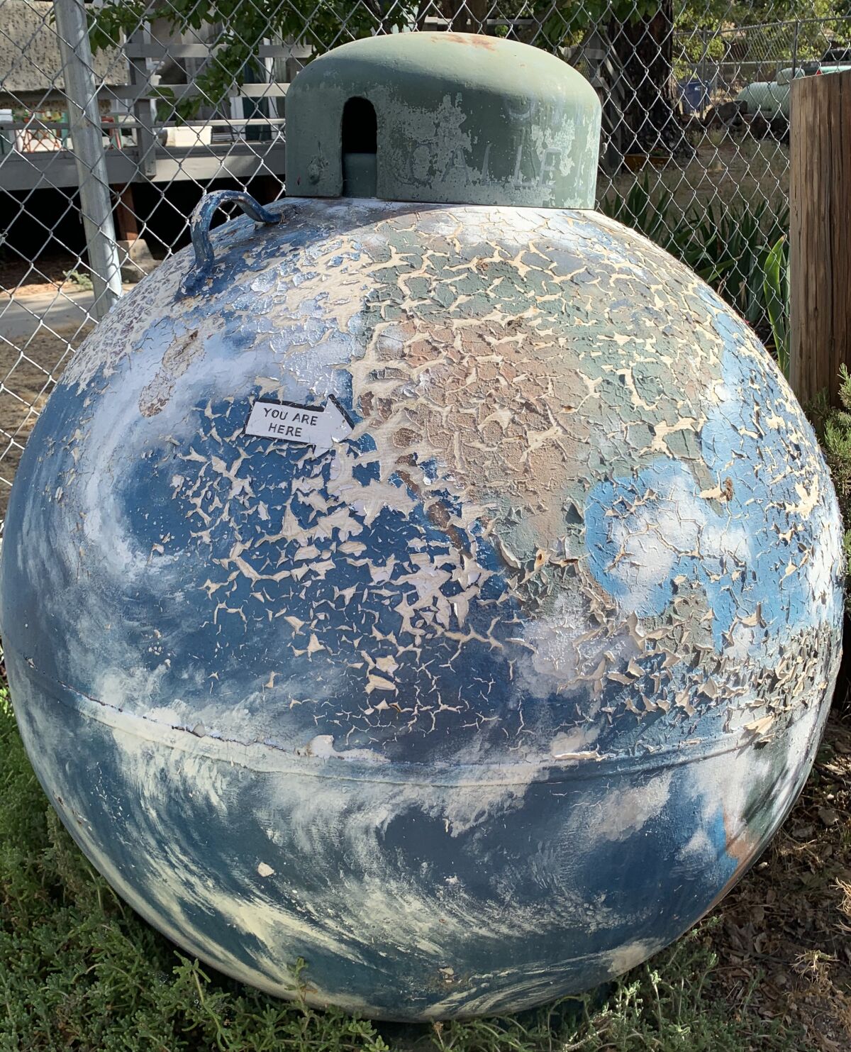 An arrow marks "You Are Here" on a spherical gas tank painted to look like Earth that is frayed and peeling.