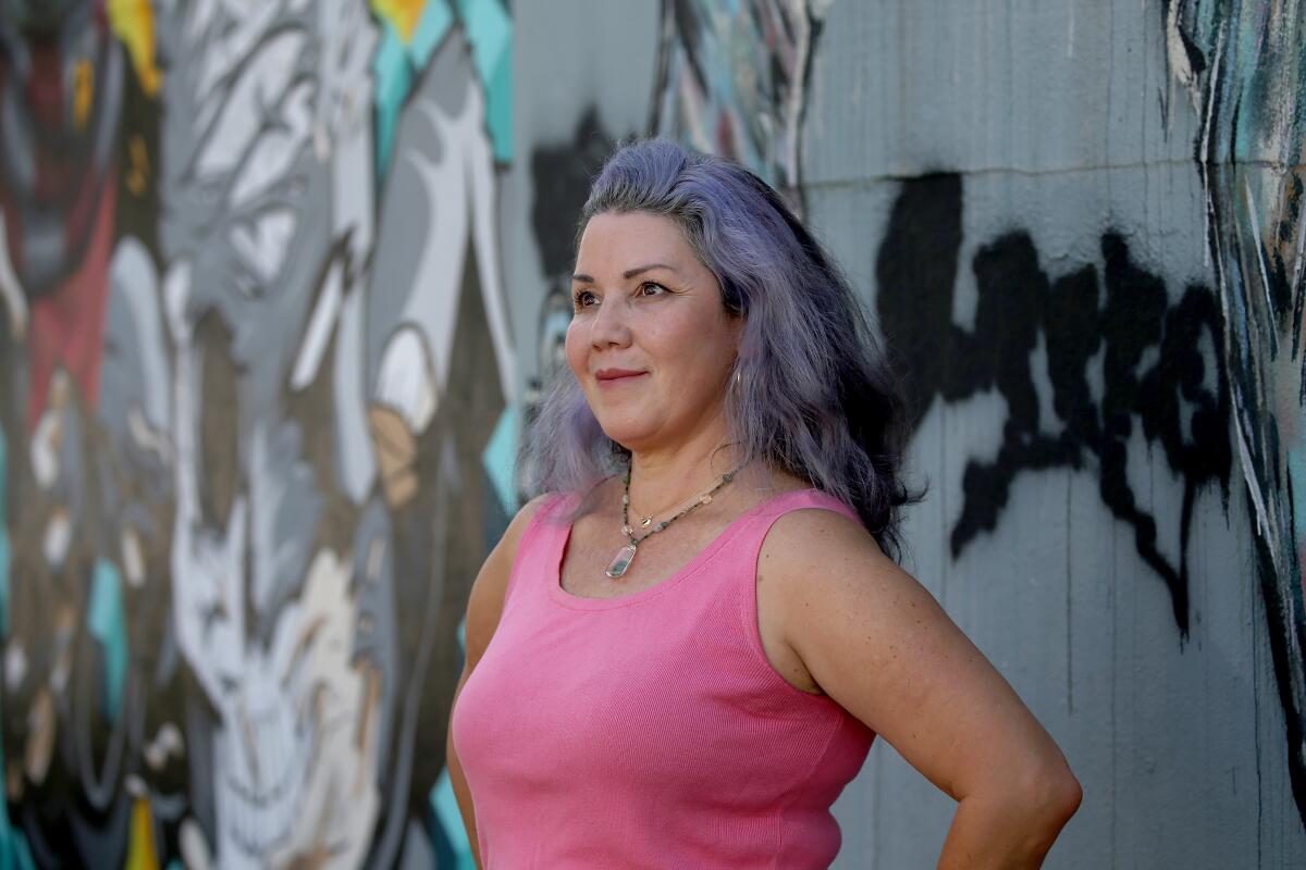 A woman with purple-tinted hair and a pink tank top poses in front of a graffitied wall.