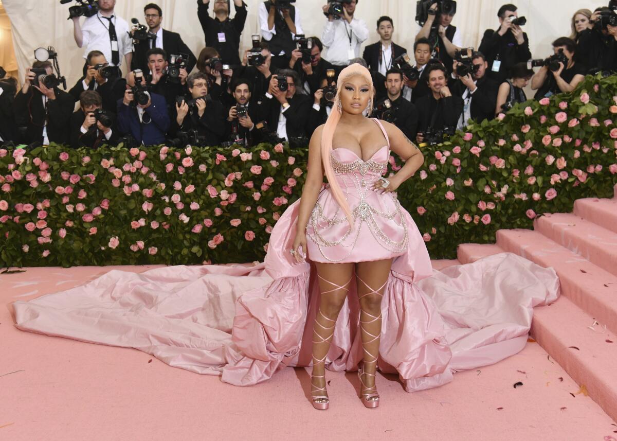 Nicki Minaj stands in a short pink gown with a long train in front of pink and green foliage and photographers.