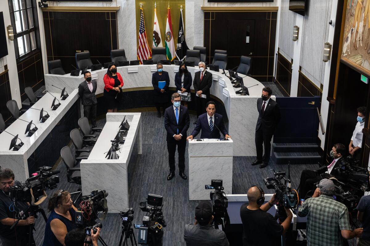 An overhead view of two men standing at a podium in government chambers, flanked by officials and news photographers.