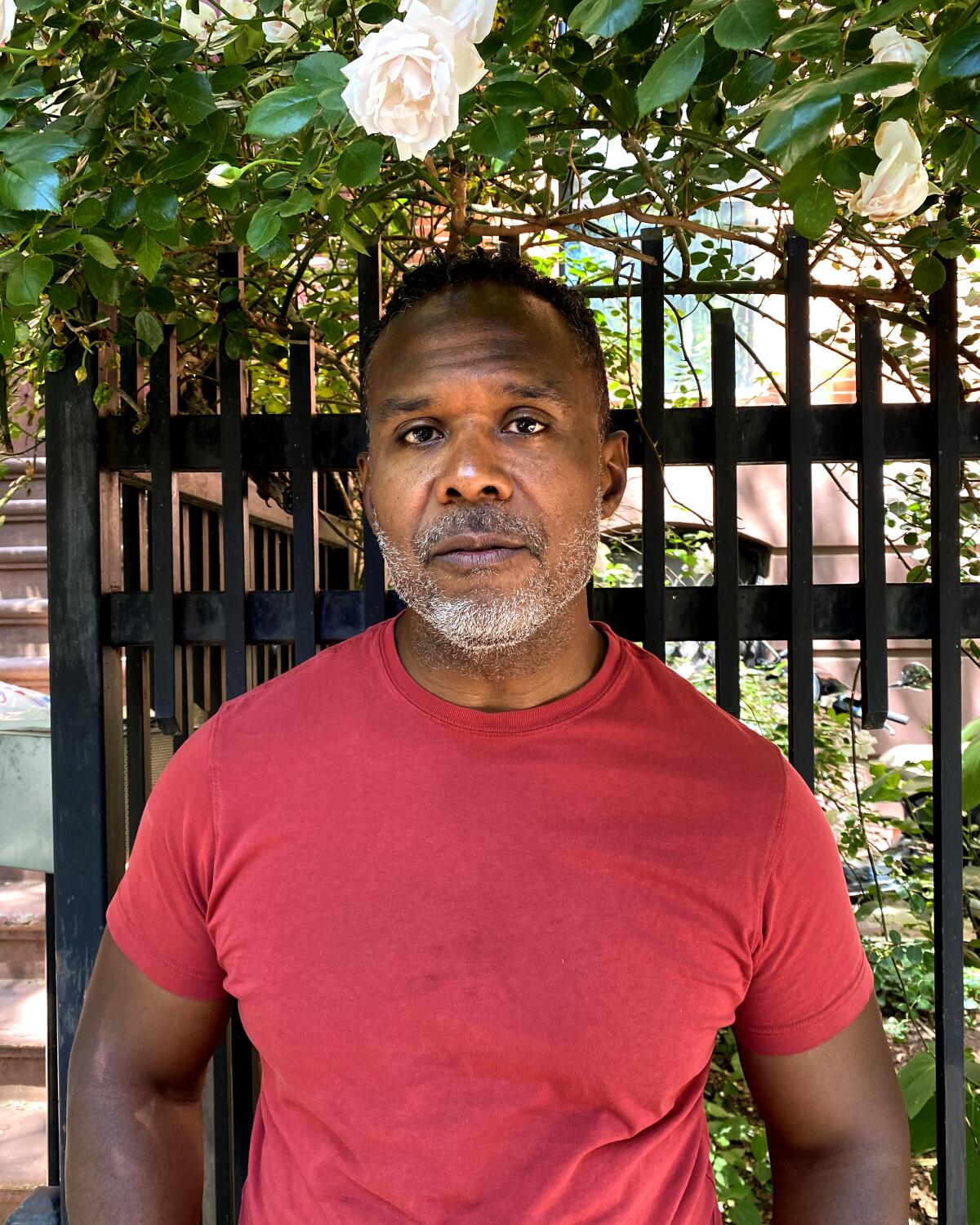 A man in a red T-shirt stands outside, a fenced-in garden behind him.