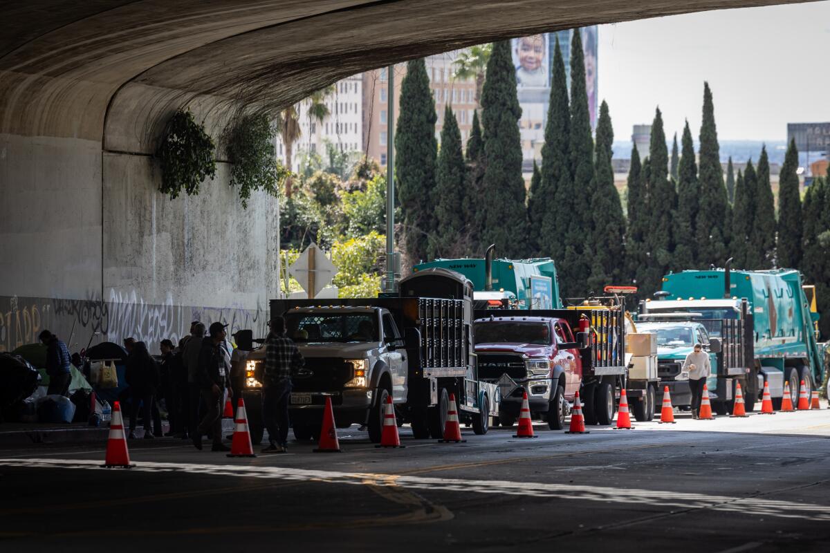Trucks and orange cones on a street under a freeway overpass.
