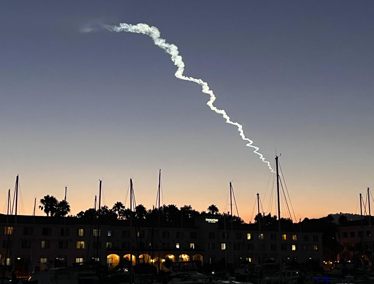 A exhaust trail rises over the masts of moored sailboats. 