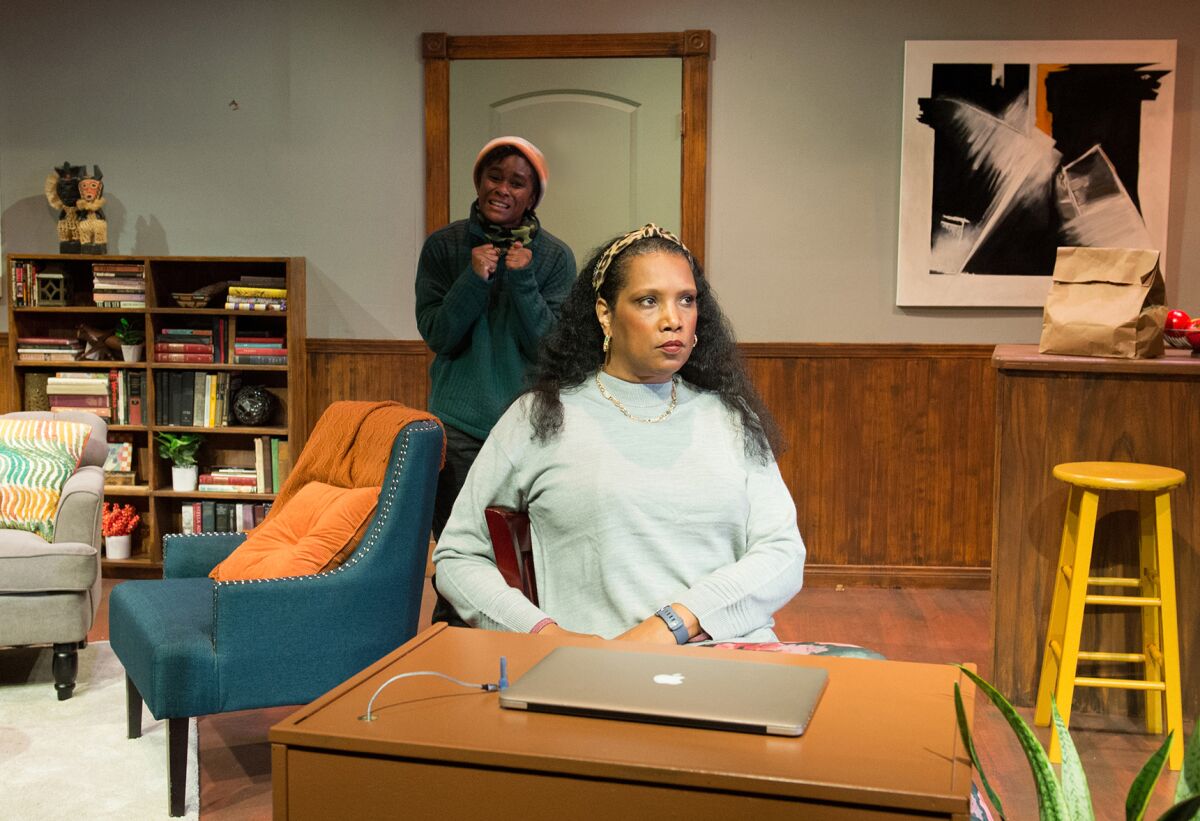 Two women on a living-room stage set, with chairs, bookcase and table with laptop computer.