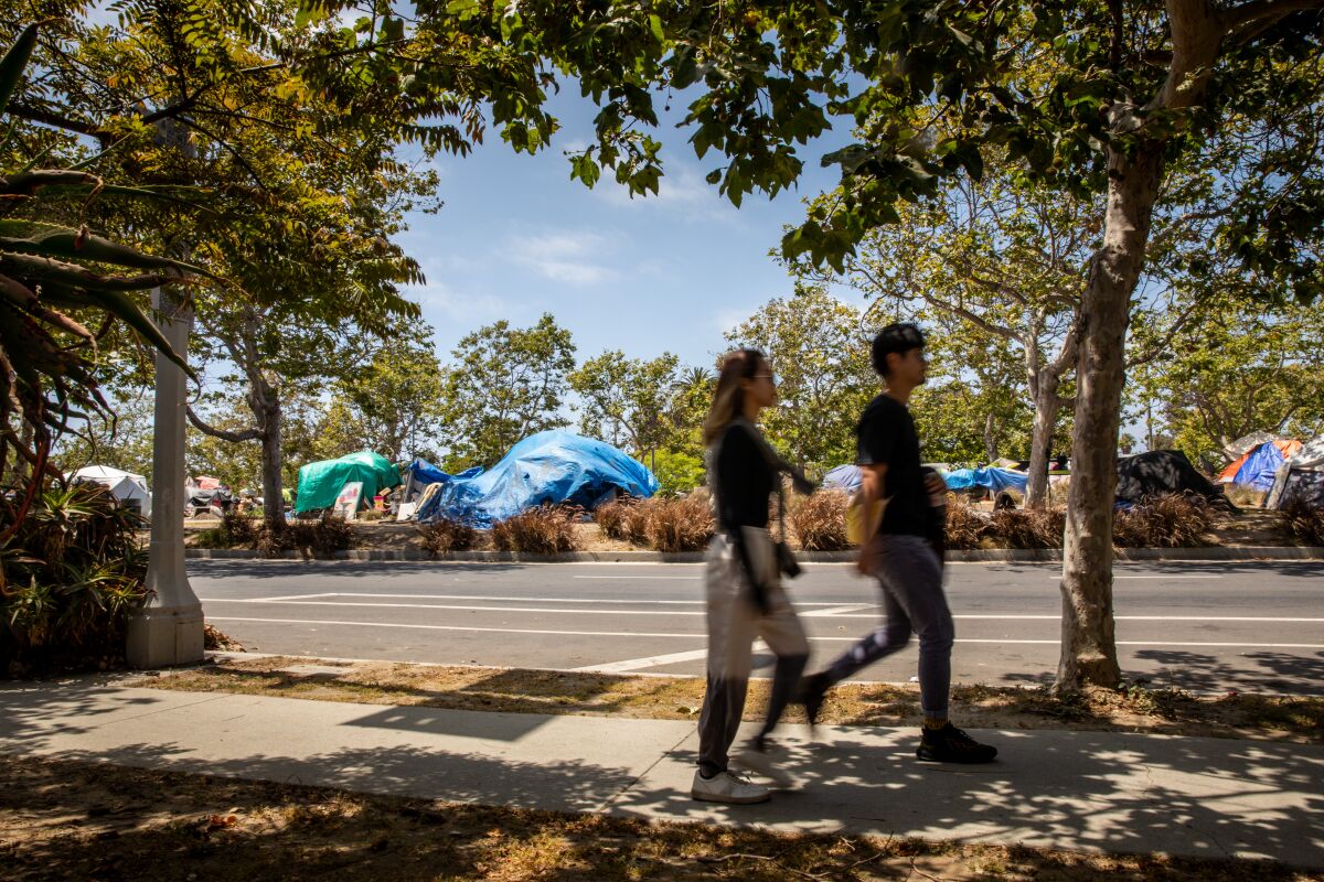 People walk along South Venice Boulevard, with tents and various structures seen behind them.