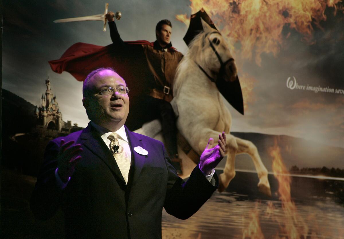 A man in a suit stands in front of an image of a man riding a horse and wielding a sword amid flames.
