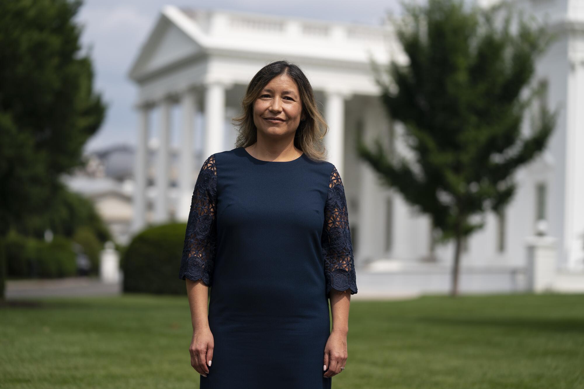 A woman with brown hair, in a navy blue dress with elbow-length sleeves, stands in front of the White House