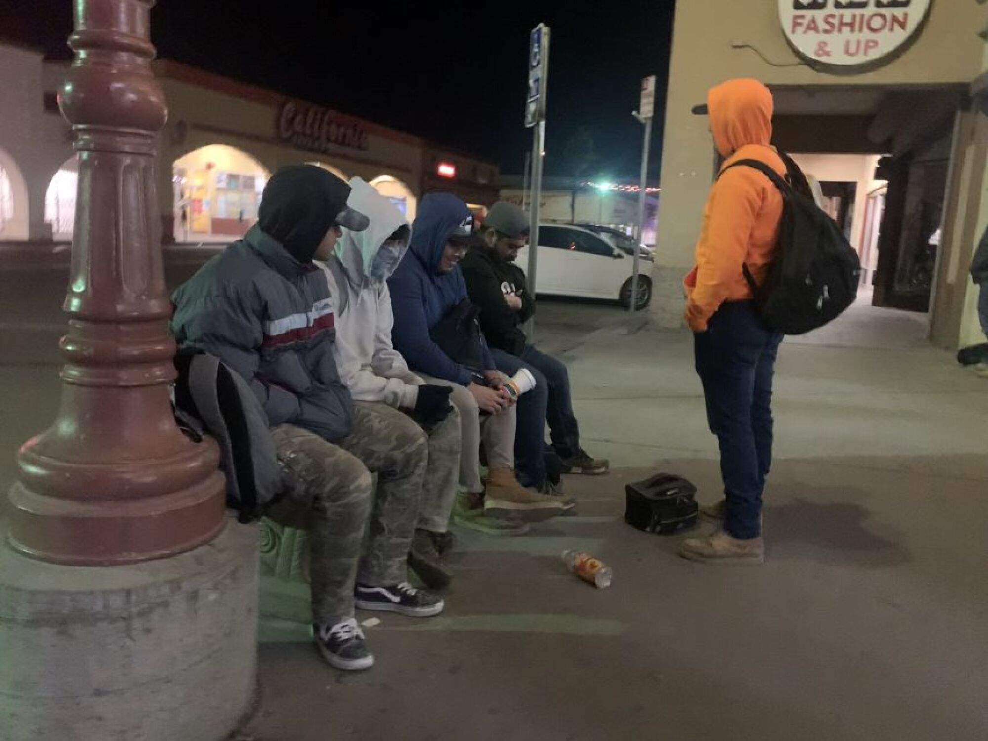 Four men in hoodies wait in a dark shopping center parking lot
