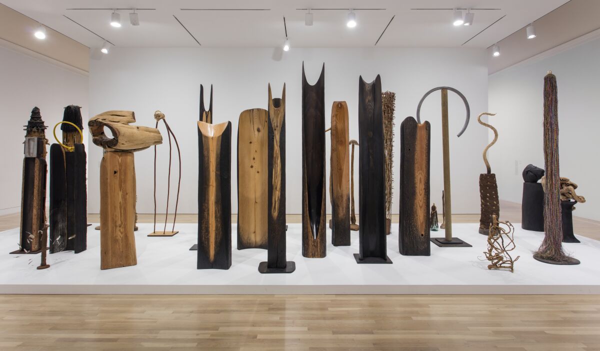 A collection of abstracted wooden totems — looking like spirits — rest on a broad, shallow museum plinth.