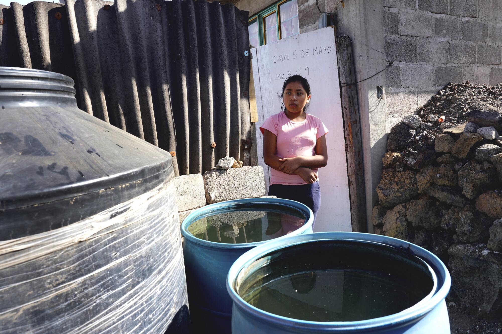 A young woman in a pink top stands by barrels of water. 