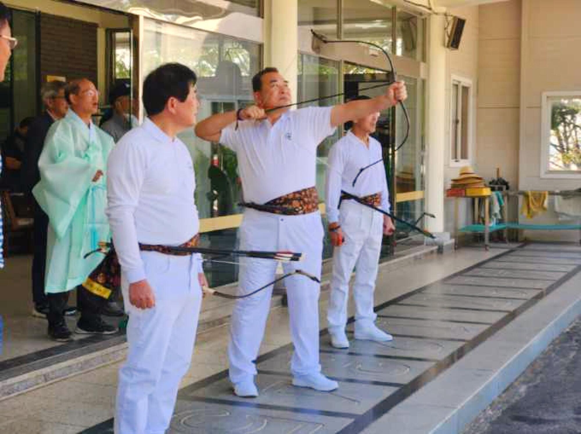 Three men wearing all white stand in a row holding bows and arrows 