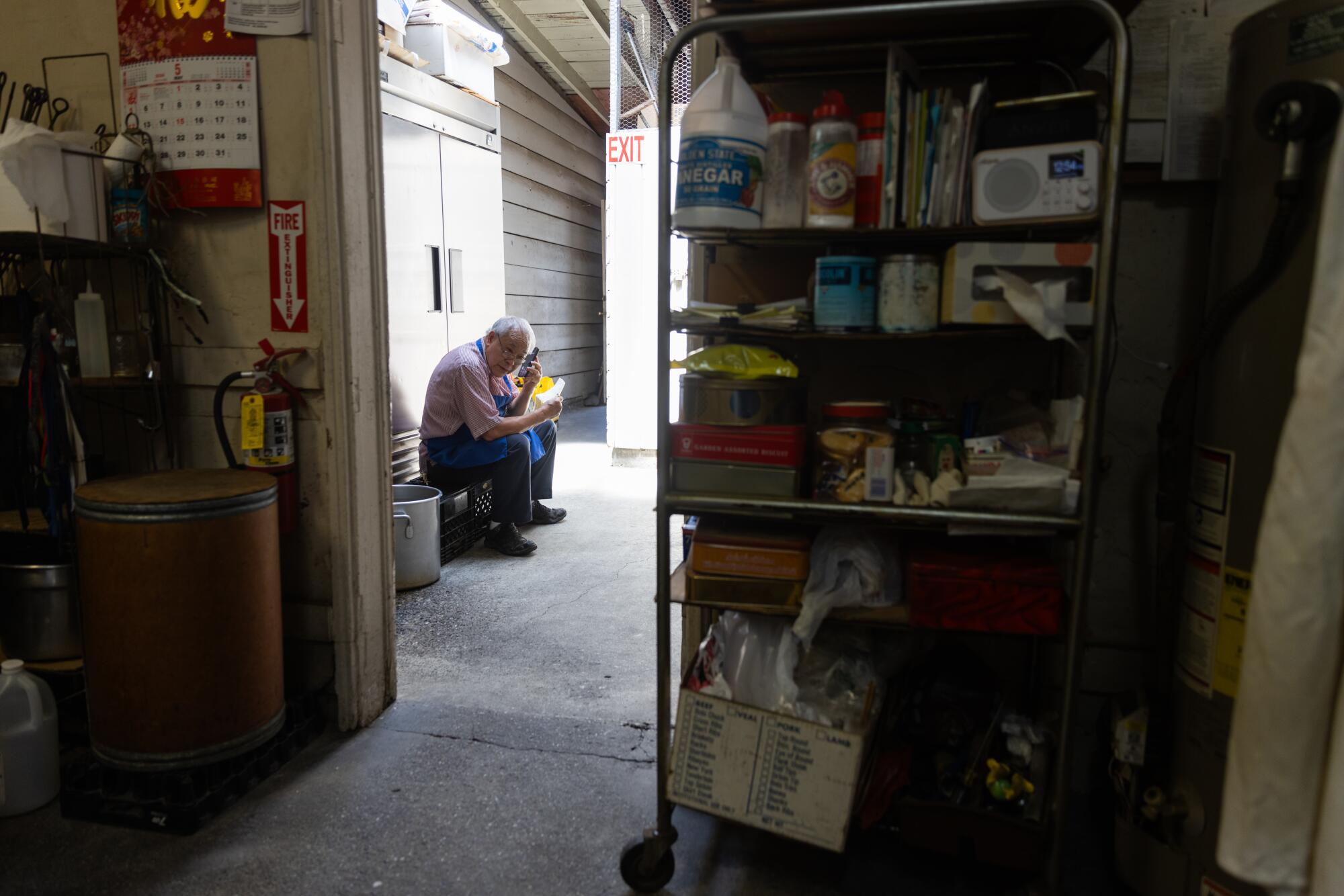 A man sits on a small chest while on his cell phone outside a restaurant kitchen.