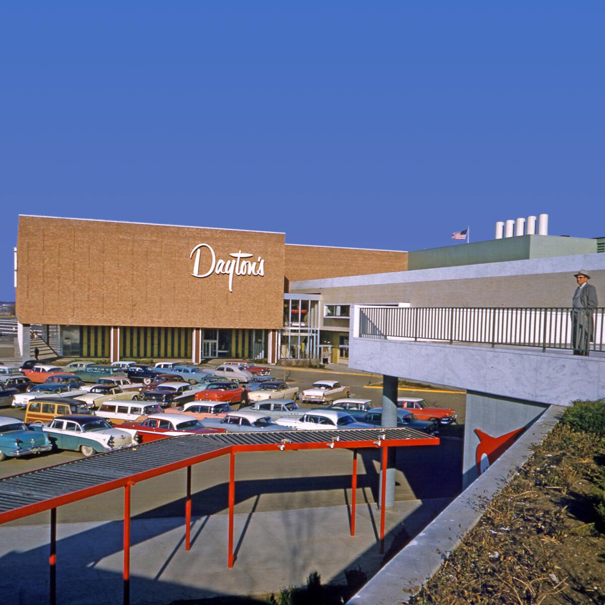 A '50s photo shows a man in a grey suit and fedora looking at a Dayton's department store from an elevated platform