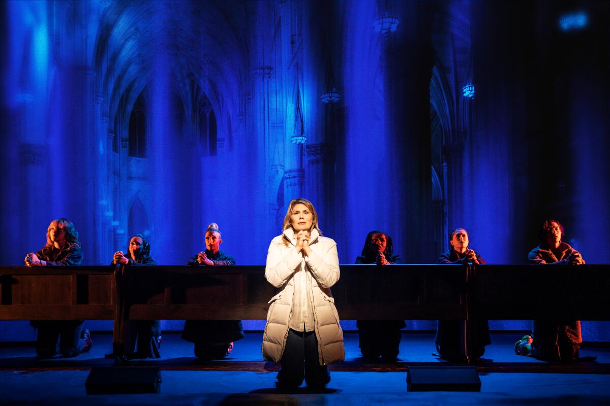 A woman in a down coat kneels in a spotlight, with a row of actors kneeling behind her.