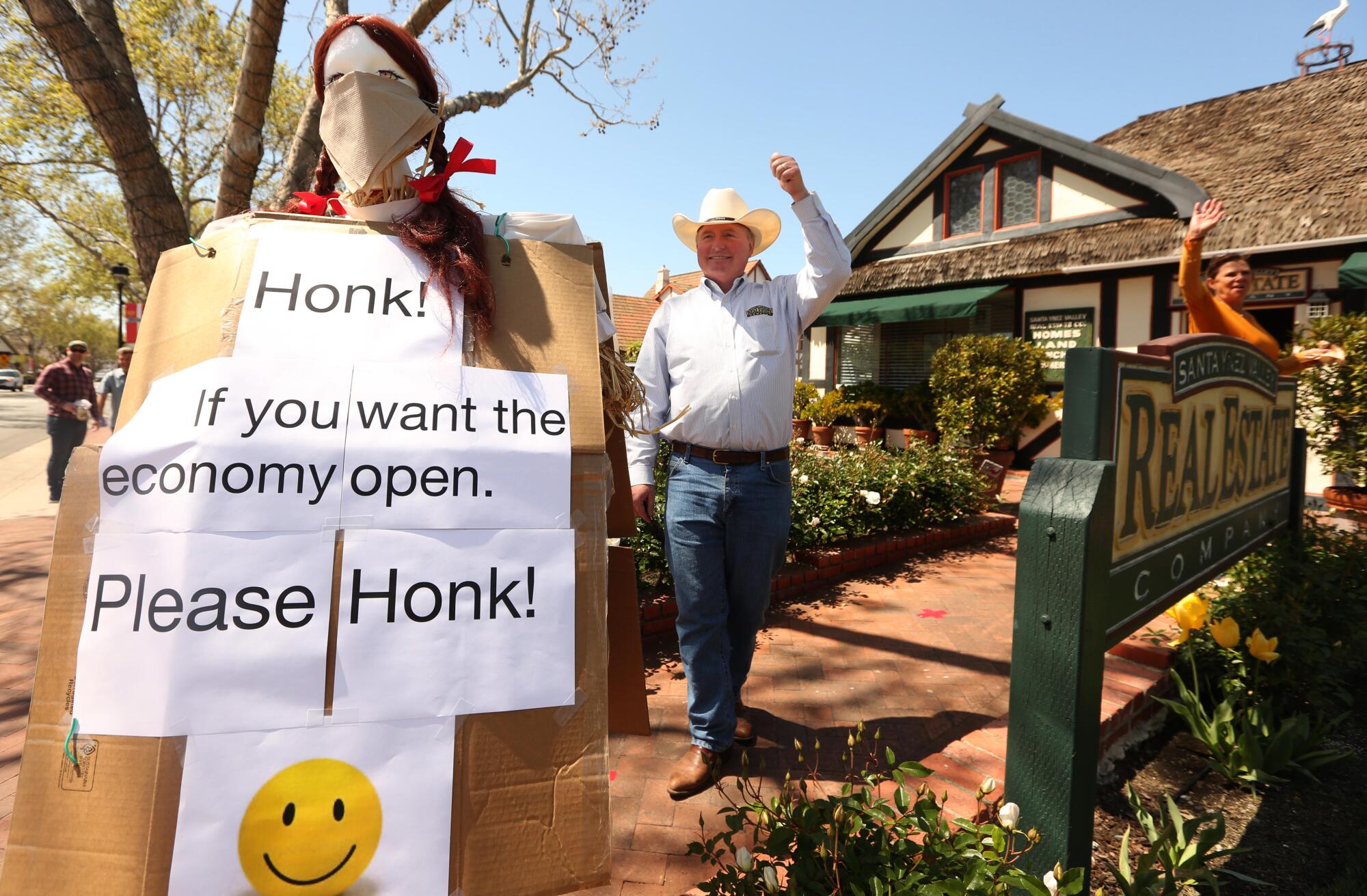 Realtor Allan Jones and his wife acknowledge a honking car last week while standing next to a sign he erected at his real estate office in Solvang in hopes the economy opens up again.