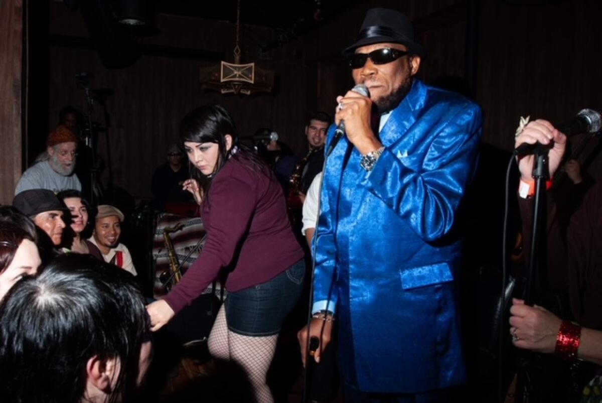 An older man wearing a shiny blue suit, sunglasses and a black hat sings in a club.