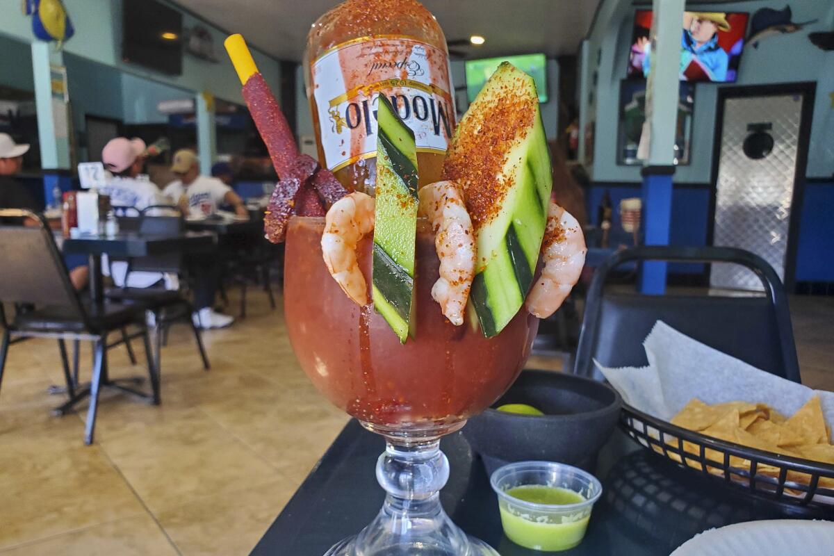 A glass goblet on a table, its rim full of shrimp and sliced cucumber, with an upside-down Modelo bottle in it.