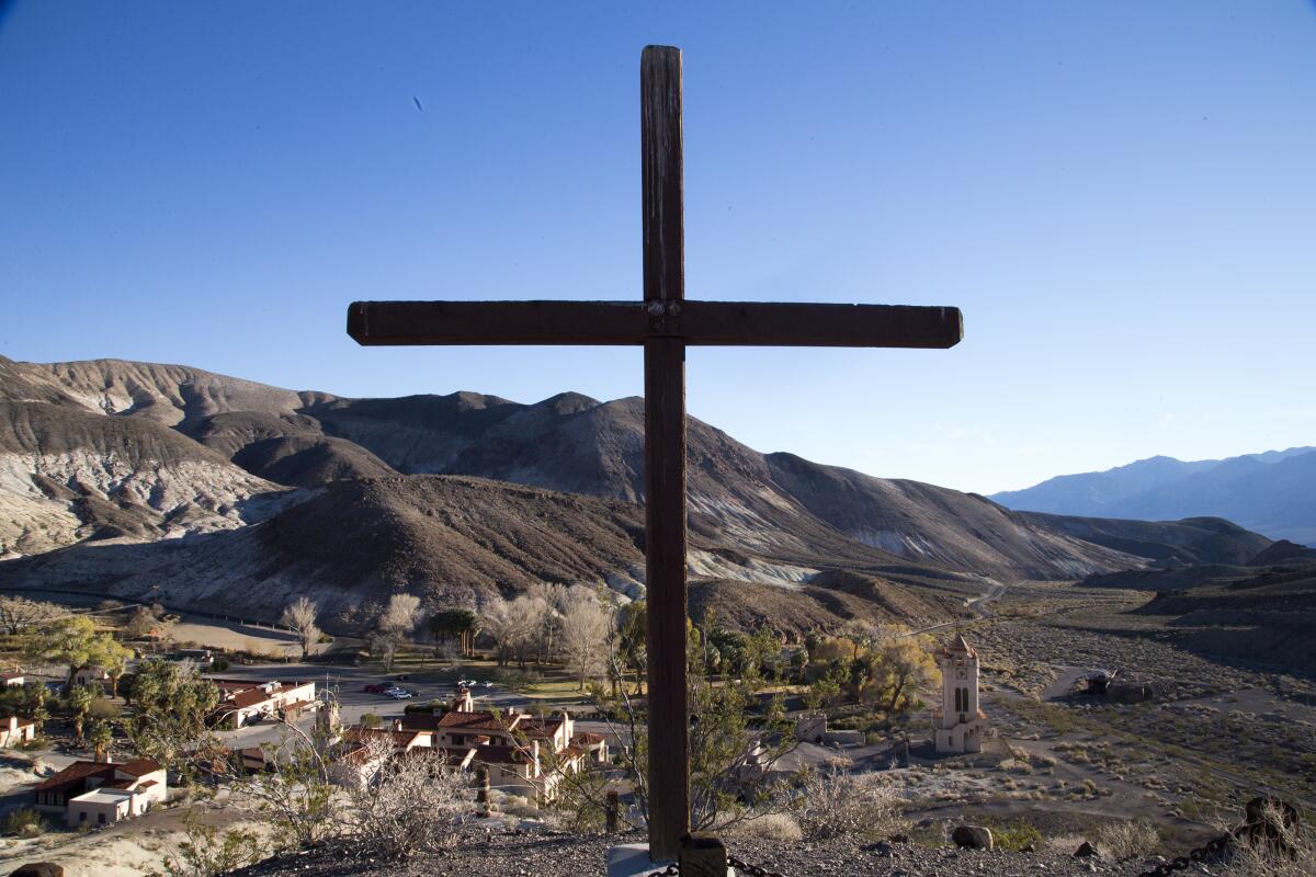 A wooden cross rises in the foreground with homes and mountains in the background.