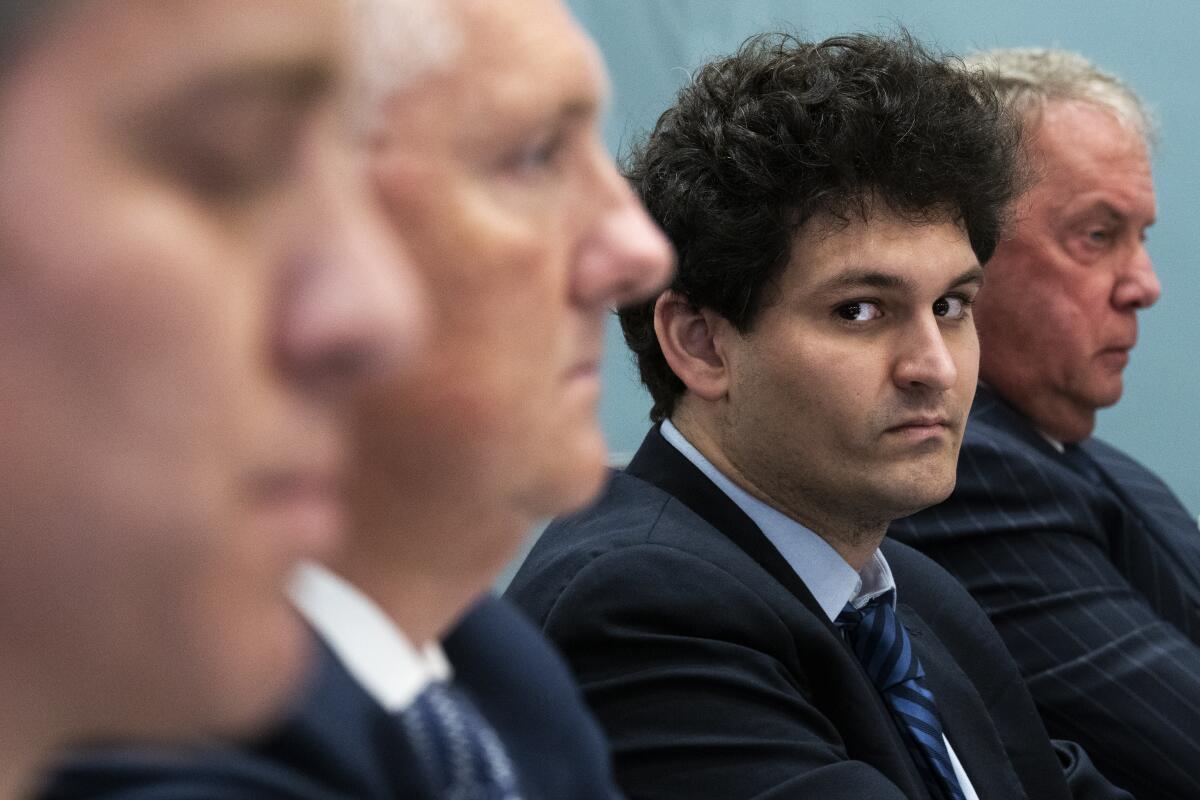 A man with shaggy hair and a navy blue jacket looks toward the camera while three other men look ahead, grim-faced