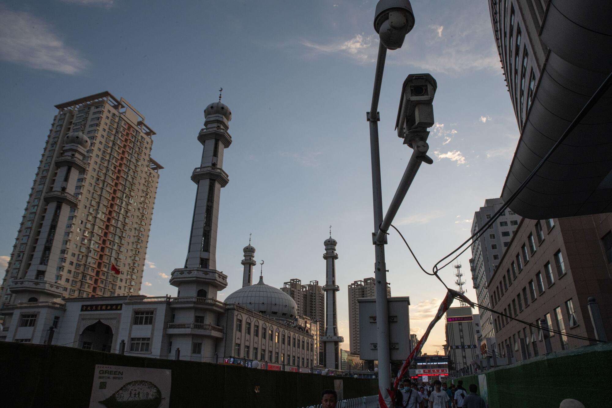 A camera mounted on a pole is aimed at a mosque with a dome and four minarets that sits amid other tall buildings 