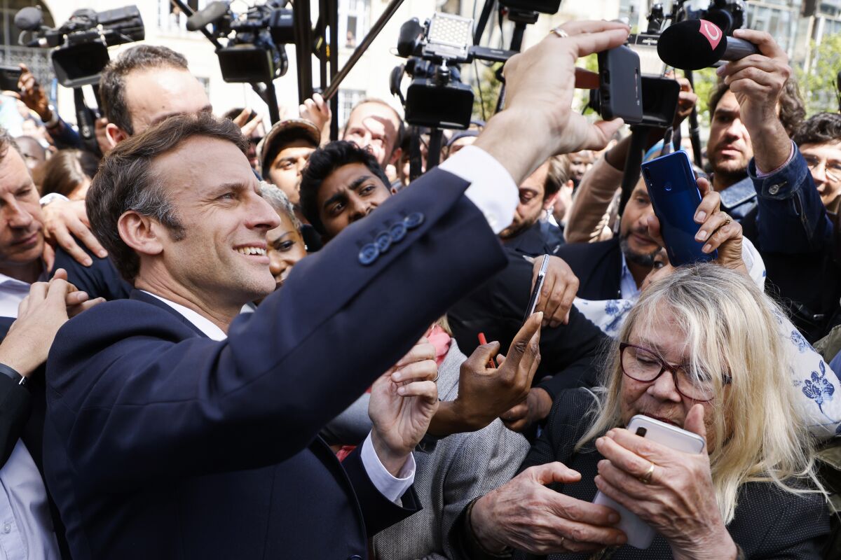 French President Emmanuel Macron smiles and holds up a phone while surrounded by cameras