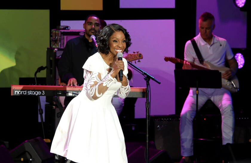 A woman in a white dress sings into a microphone onstage, with other musicians behind her.