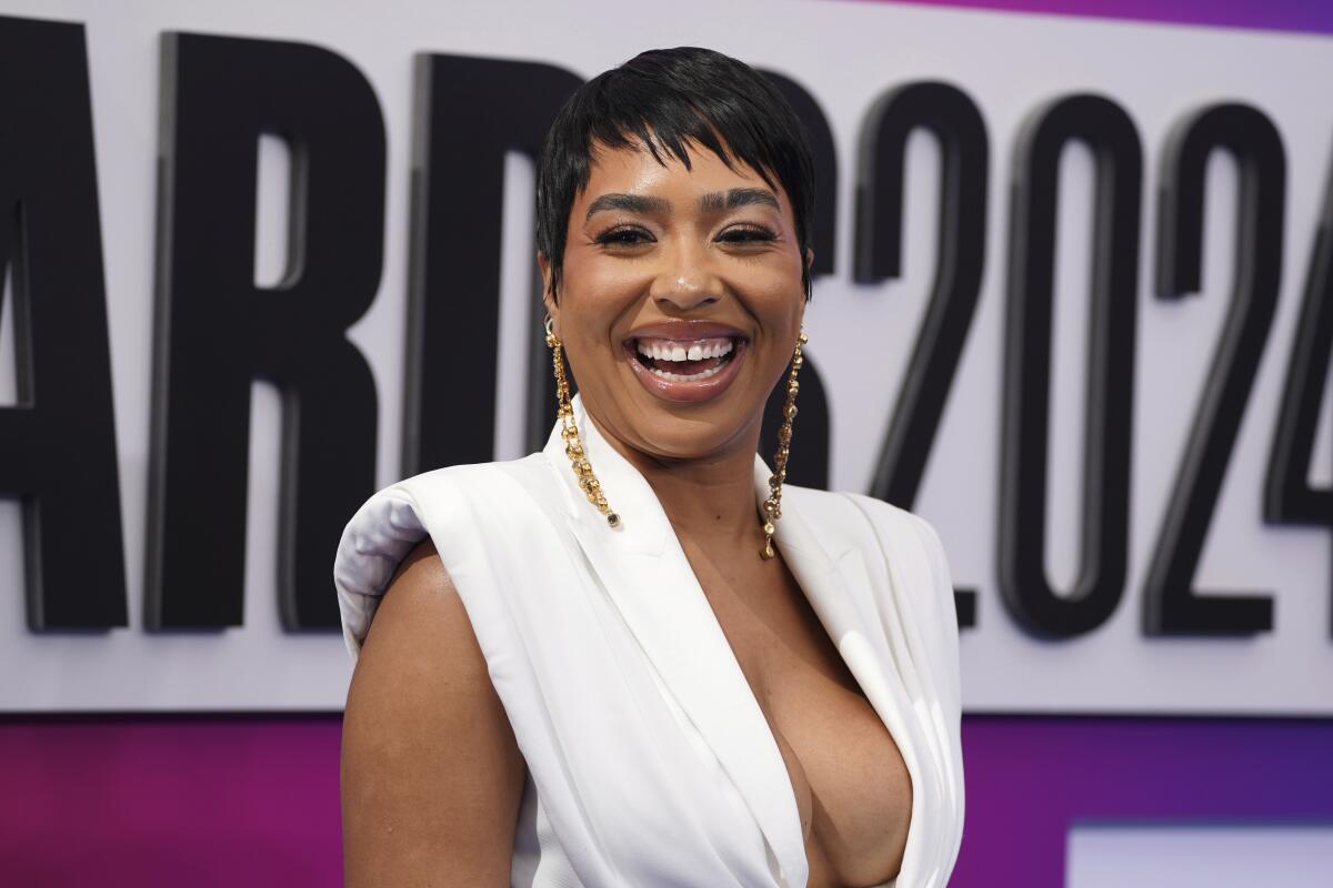 B Simone in a low-cut white dress and dangling earrings smiling against a white backdrop with black text
