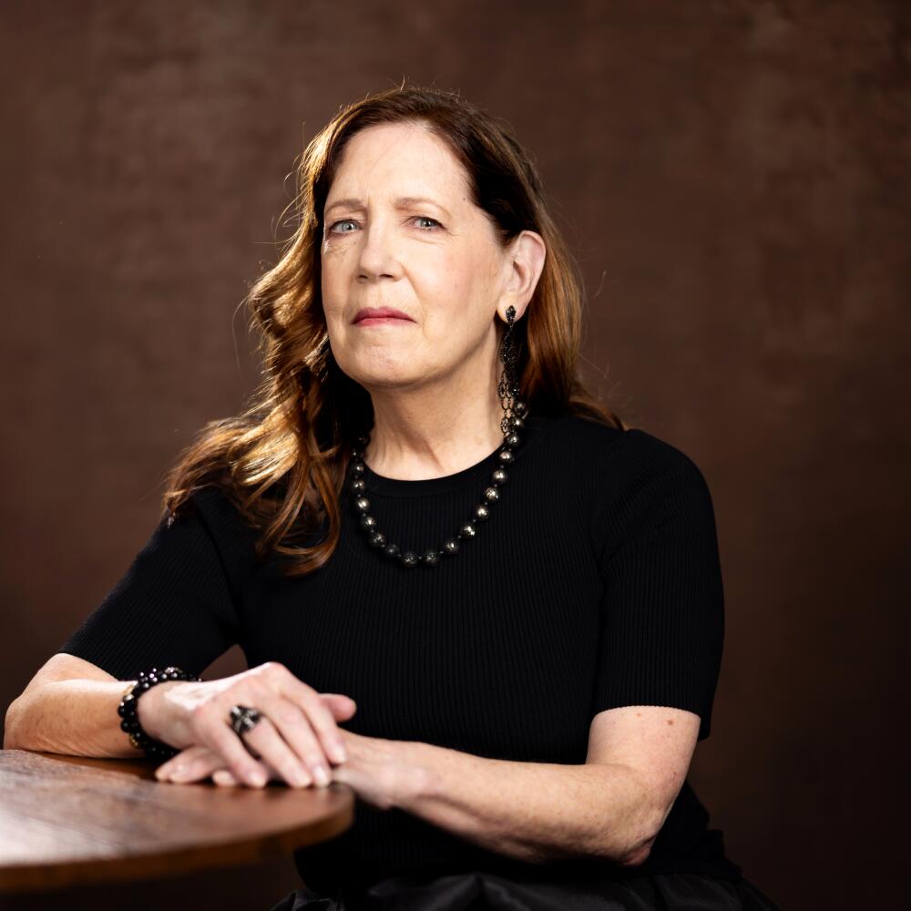 A woman with long brown hair in a black top and a black pearl necklace rests her hands on table top.