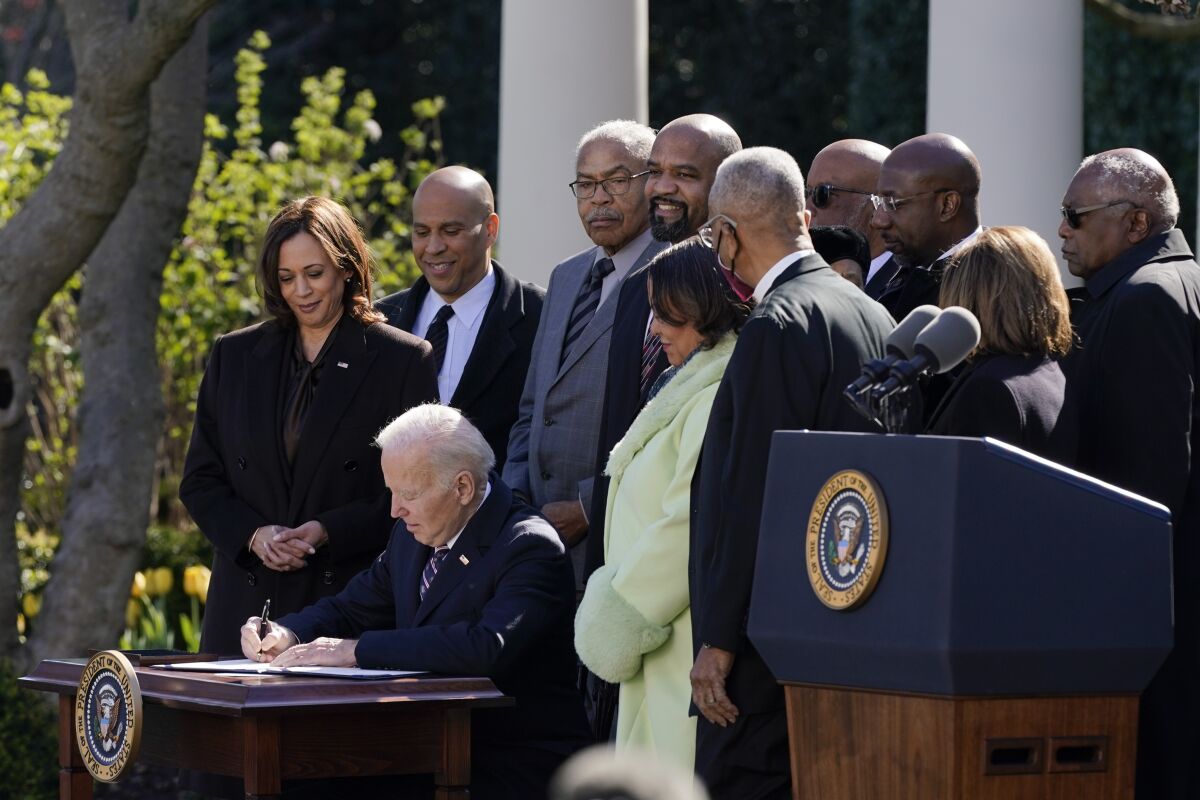 President  Biden, surrounded by onlookers including Vice President Kamala Harris, sits and signs a paper  