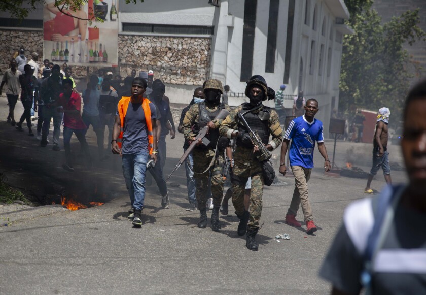 Heavily armed police in combat gear walk among civilians on a street where small fires burn